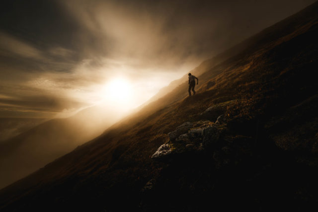 Silhouette d&rsquo;un homme marchant sur une pente herbeuse des Lofoten, &eacute;clair&eacute;e par une lumi&egrave;re rasante de fin de journ&eacute;e